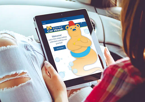 Person holding a tablet displaying a car wash app with a cartoon yellow bear wearing a red hat and blue scarf.