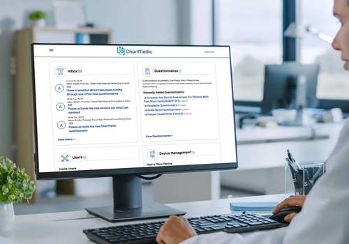 Office worker at a white desk viewing a computer monitor displaying a ChartMedic dashboard with Inbox and Questionnaires sections.