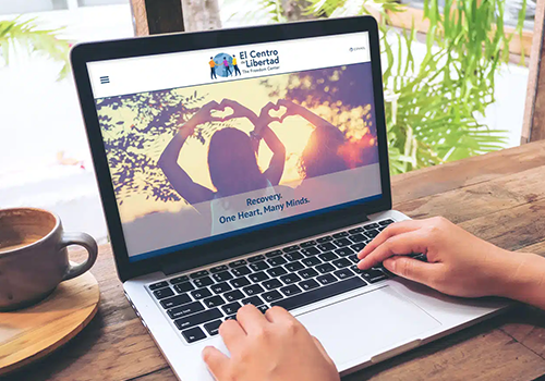 Person typing on a laptop at a wooden table, viewing the El Centro Libertad homepage with a heart-shaped hands banner on screen