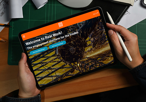 Person holding a tablet showing a job platform homepage with orange top bar and sparks background text: 'Welcome to Real Work! The employment platform for the trades'