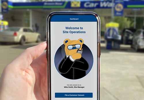Hand holding a smartphone displaying the Site Operations dashboard with a bear mascot, at a car wash backdrop