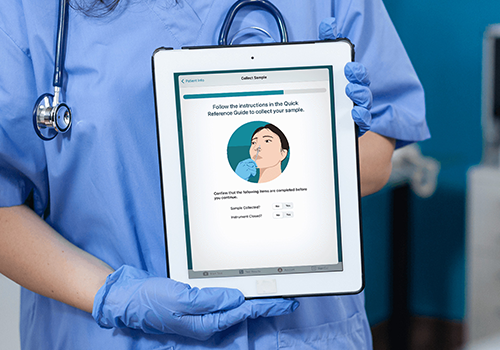 Healthcare worker in blue scrubs and gloves holding a tablet that shows a sample collection guide with a throat-swab illustration.