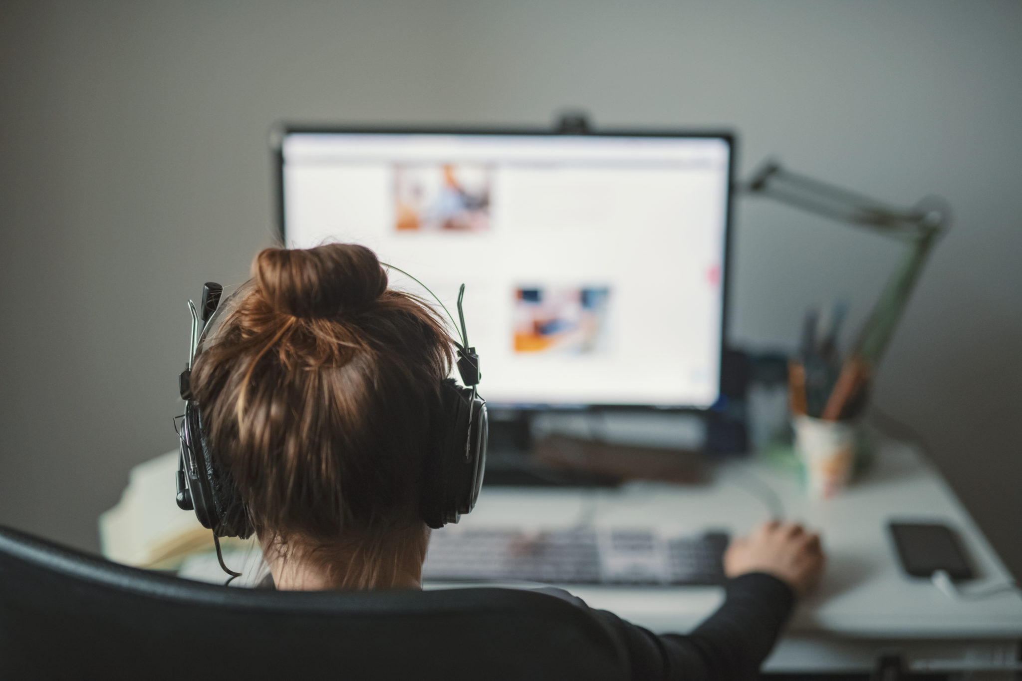 The back of a person's head with headphones and facing towards a computer screen.