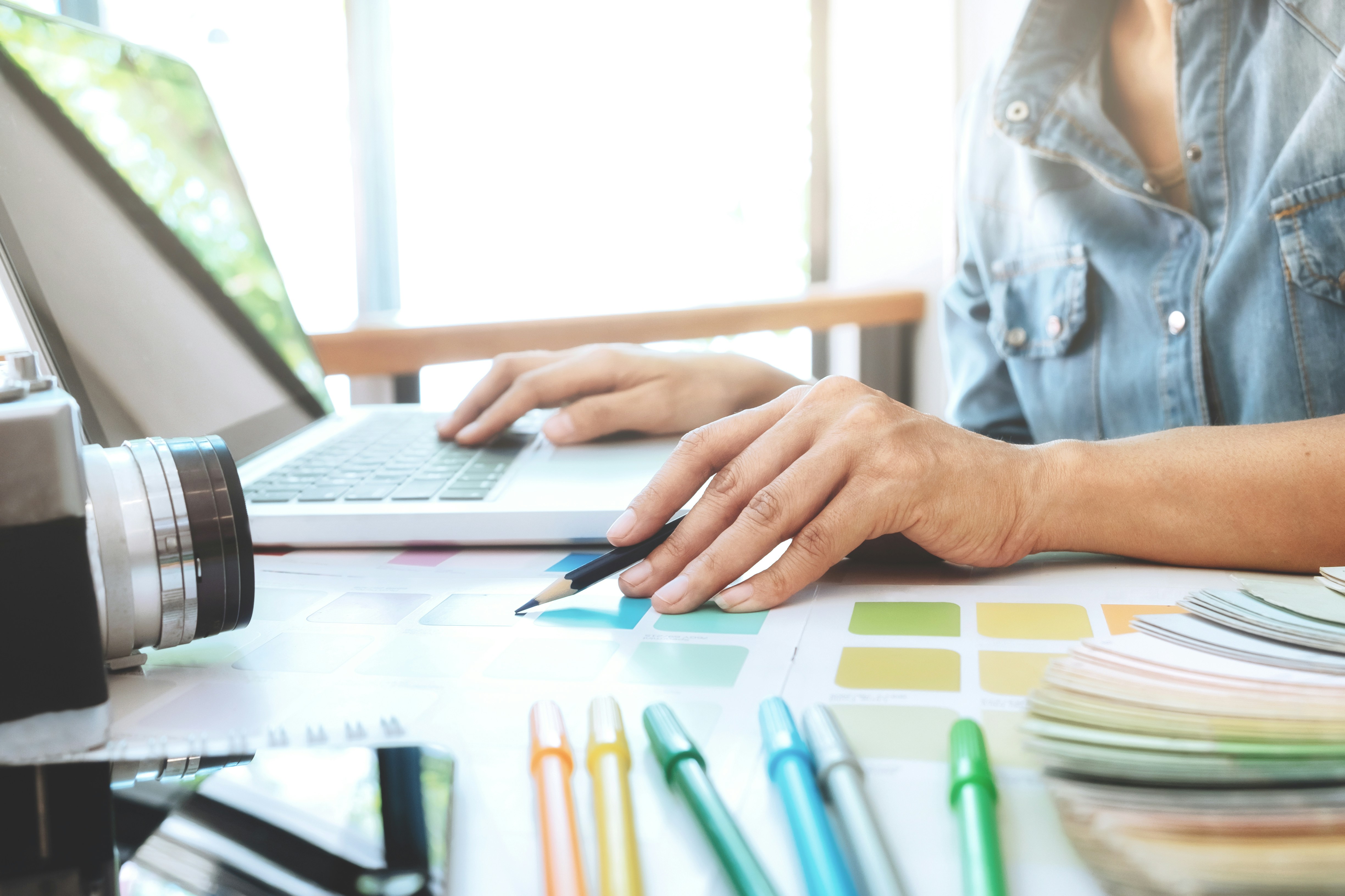 Person's hand holding a pencil with a laptop in the background and colored pencils in the foreground