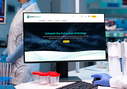 Laboratory workstation with a monitor showing a biology company website; gloved hands and test tubes in the foreground.