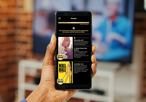Hand holding a black smartphone displaying a results page with scores and thumbnails in a dark-themed interface, blurred living room in the background.