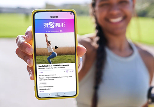 Person holding a yellow‑framed smartphone showing a sports news app (SHE SPORTS) with a video thumbnail of a person jumping on the screen.