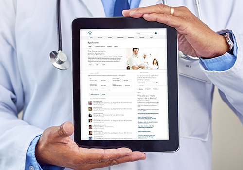 Doctor in blue scrubs holding a tablet that displays a medical website with articles and a chat feed, with a stethoscope visible around the neck.