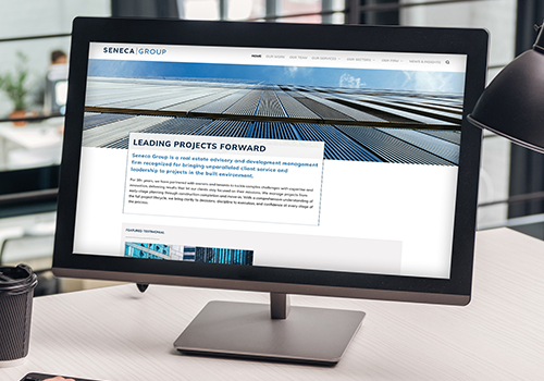 Desktop monitor on a light desk displaying a corporate website with a blue hero image and a 'Leading Projects Forward' content card.