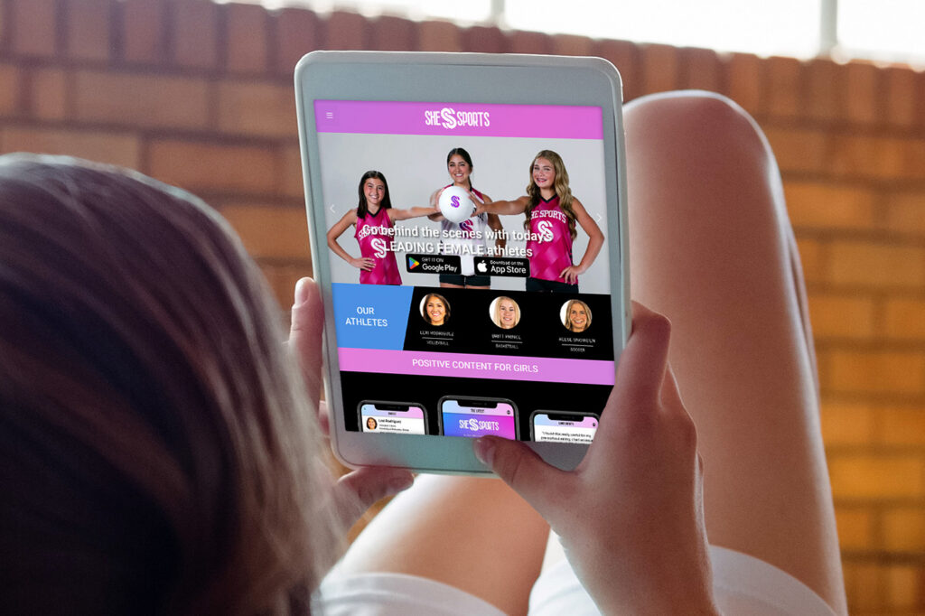 Person holds a tablet showing a pink-themed sports site for girls with three athletes on screen and app icons below.