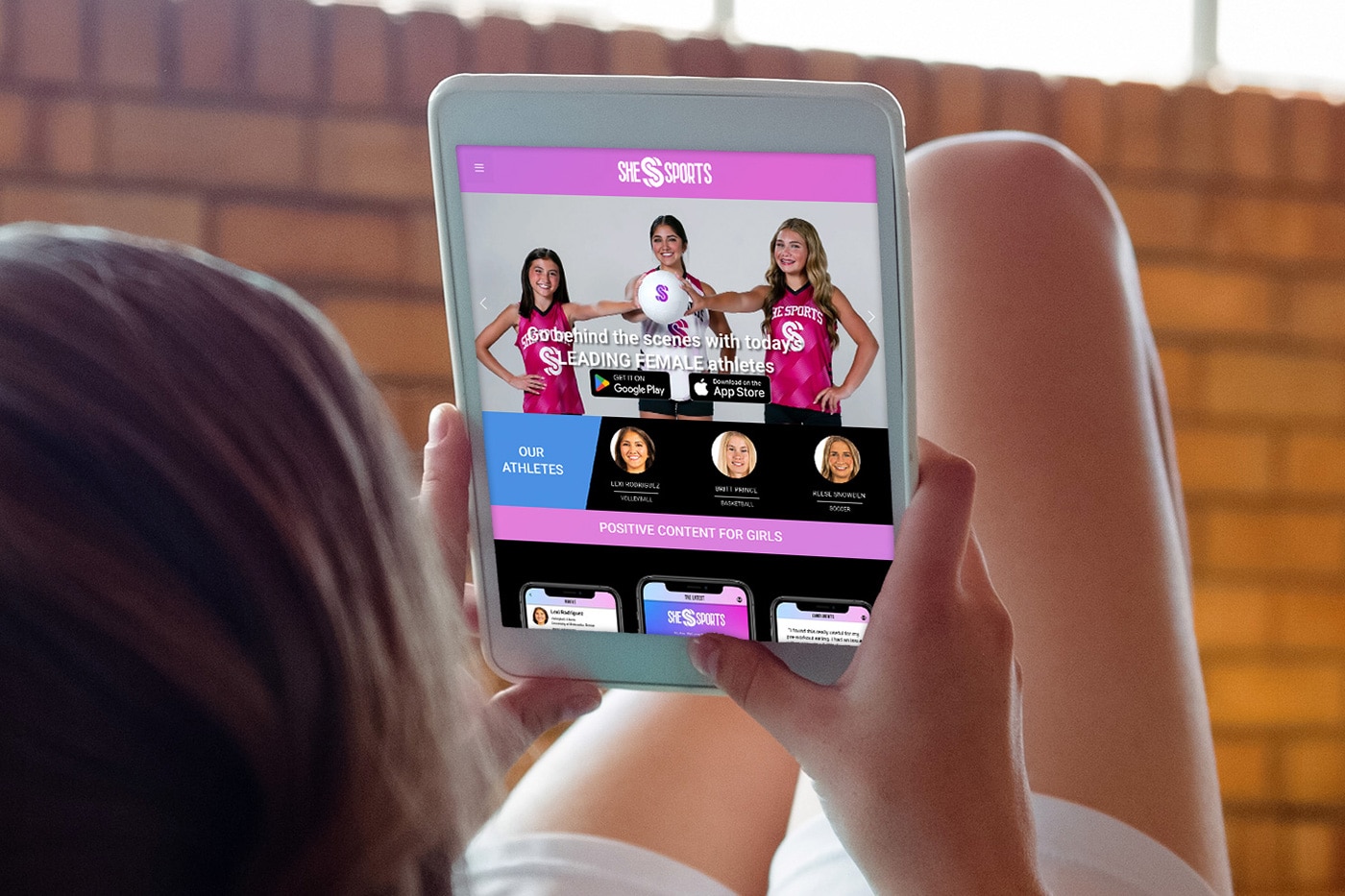 Person holds a tablet showing a pink-themed sports site for girls with three athletes on screen and app icons below.